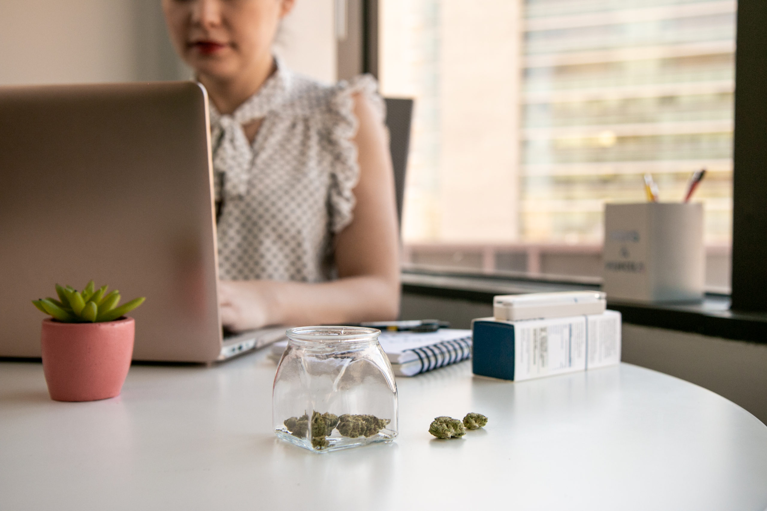 A glass jar of cannabis buds sit on a white office table in an office environment with a downtown view outside the window. A woman is working on a laptop computer with a vape pen by her side. A succulent in a pink pot adorns the desk.